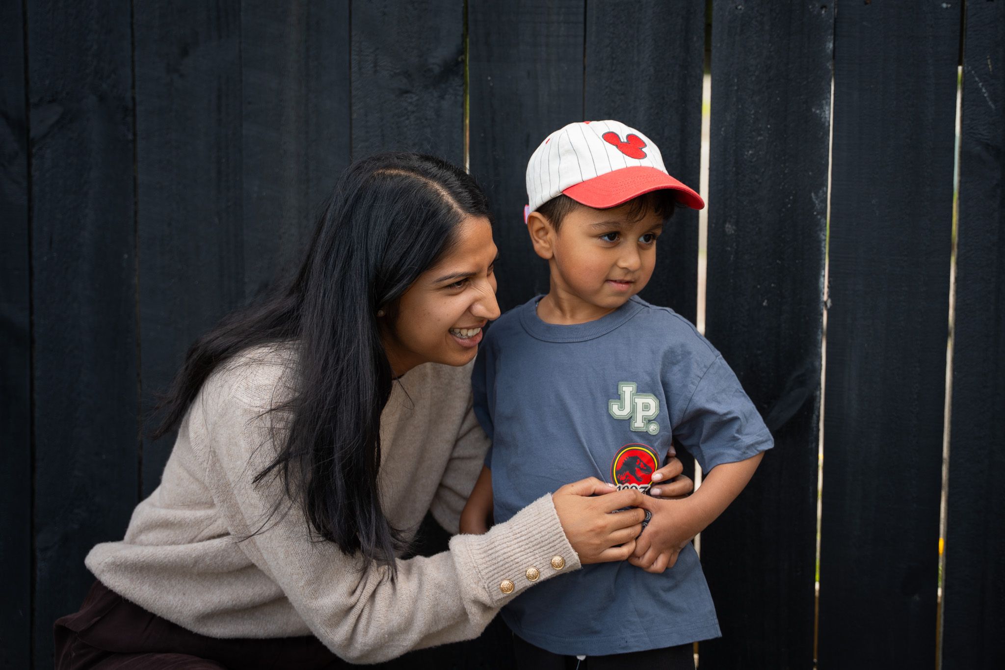 Aunty crouching and laughing beside her young nephew in a grey t-shirt and red-white cap against a dark timber fence during a candid family session in Auckland