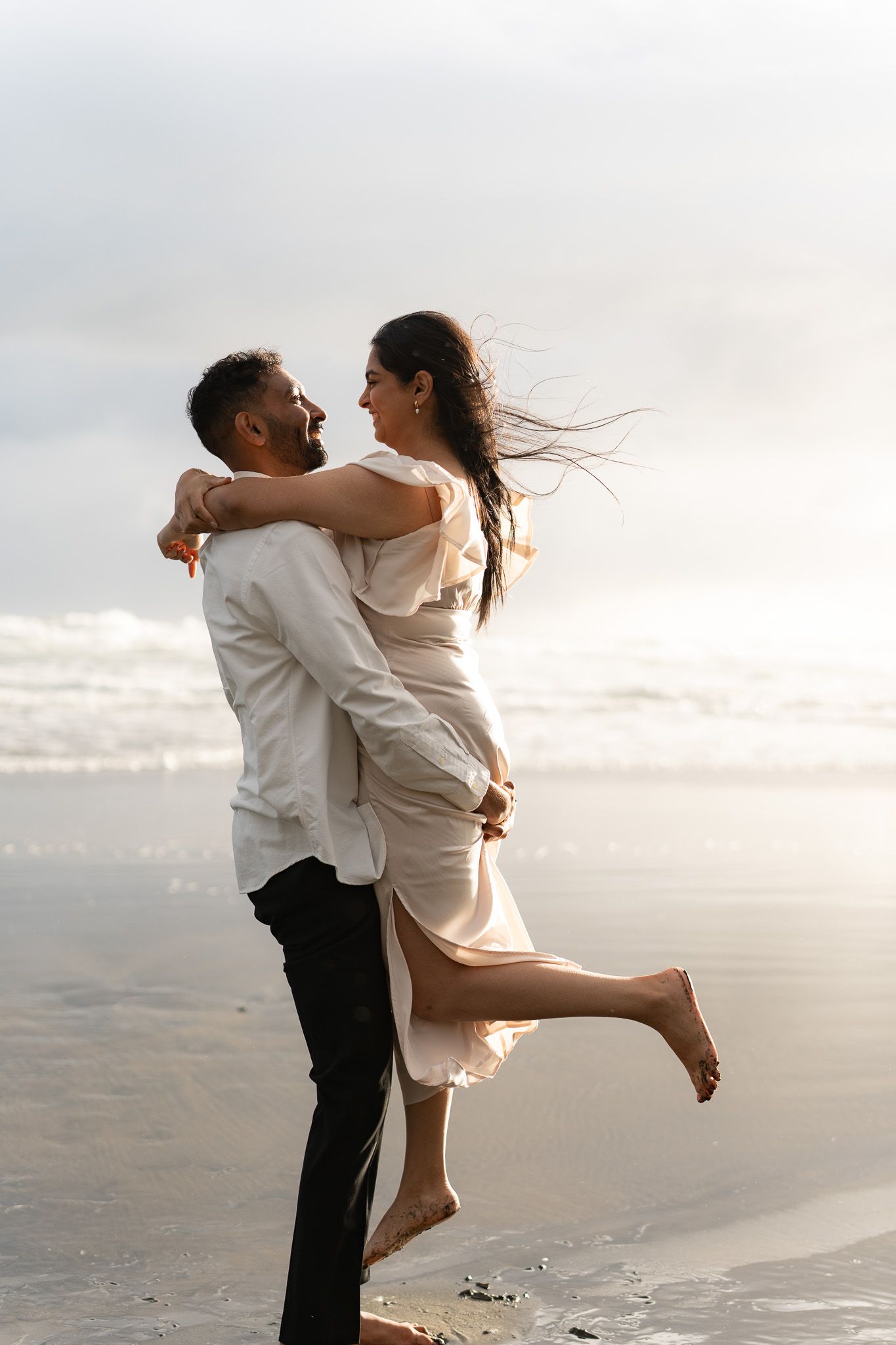 Man lifting his partner off the ground as she smiles and kicks up a bare foot on the beach