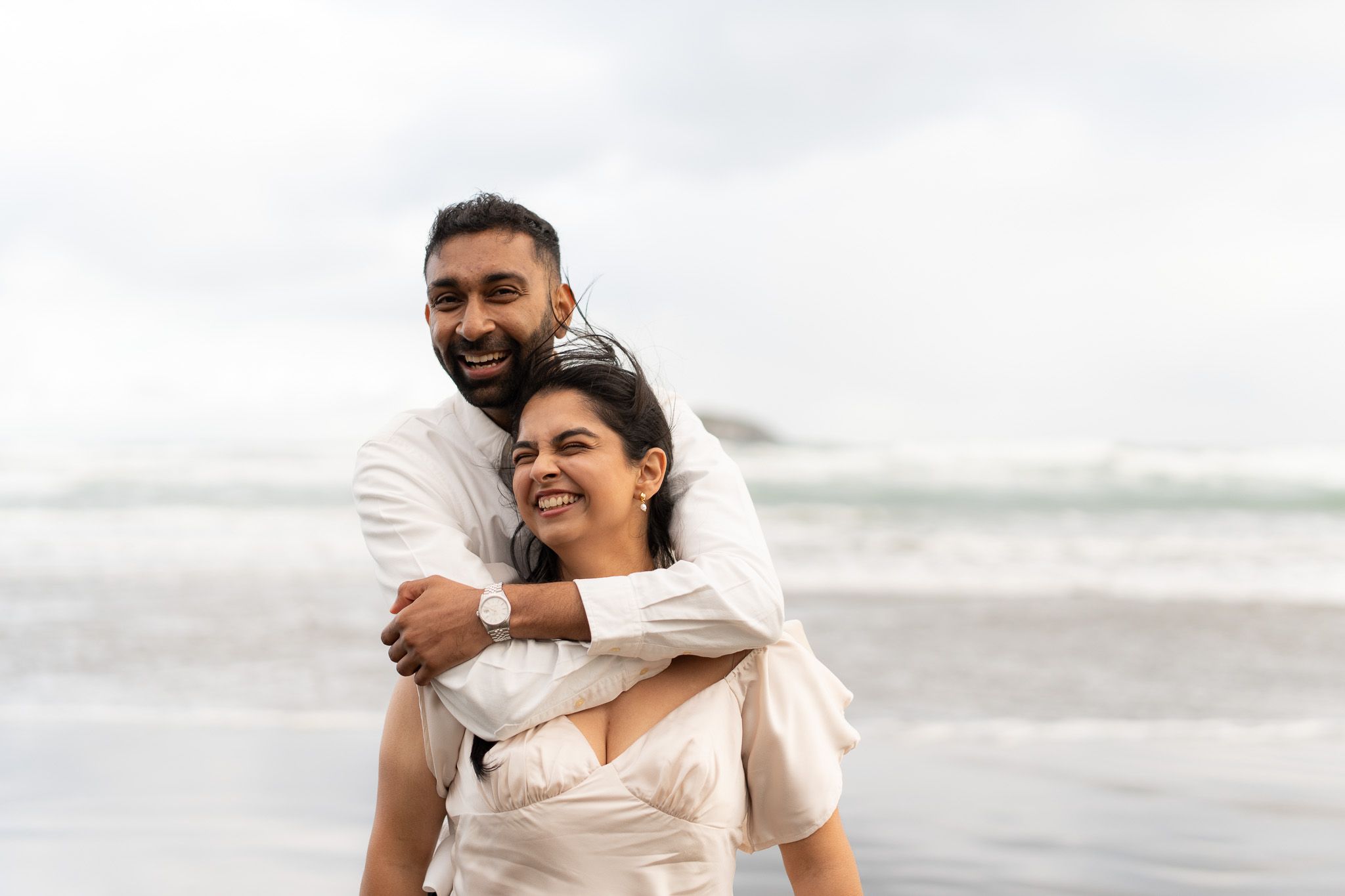 Man wrapping his arms around his laughing partner from behind on a misty beach