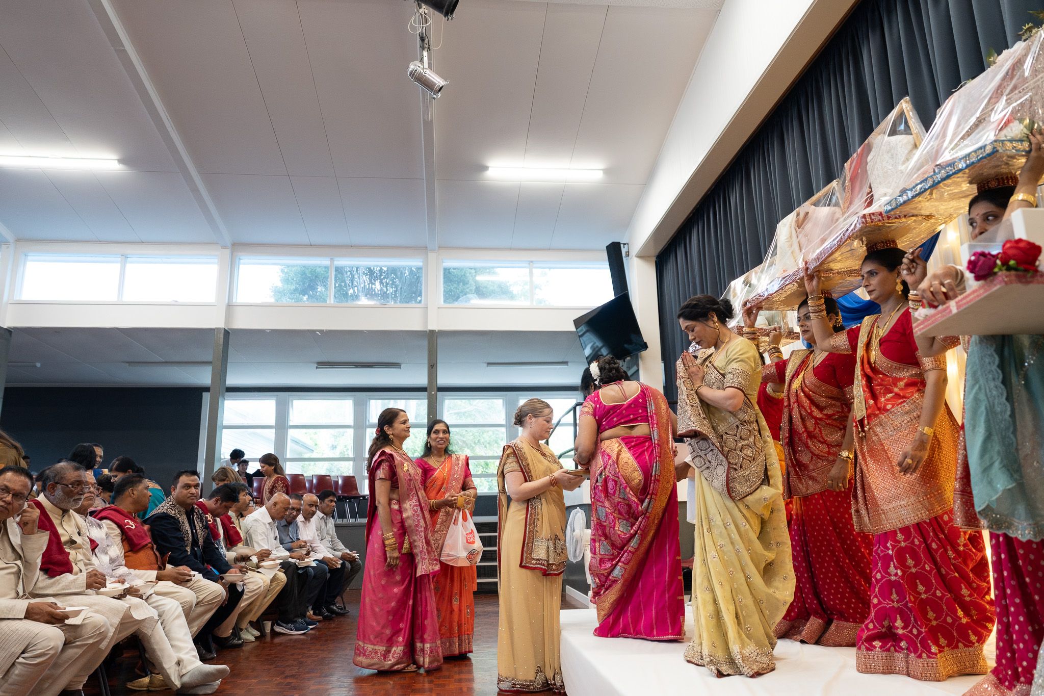 Women carrying ceremonial canopy during Indian Mosaru ceremony with family and guests seated inside hall during wedding photography in Auckland