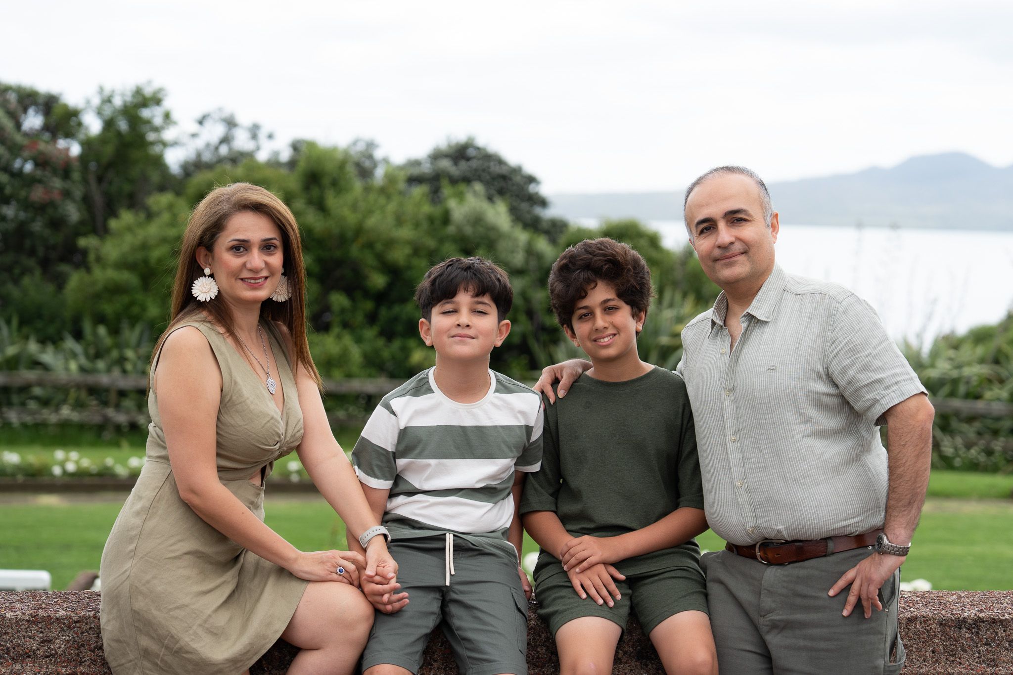Mona, Ali and their two boys sitting together with greenery and water views during a family photography session in Auckland