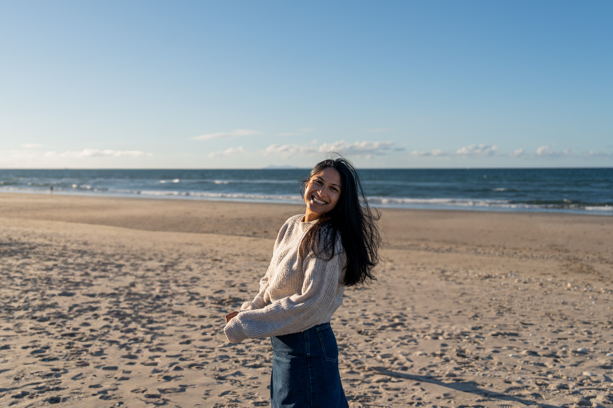 Woman smiling brightly while standing on a sandy beach with waves in the background during a relaxed outdoor portrait session in Auckland