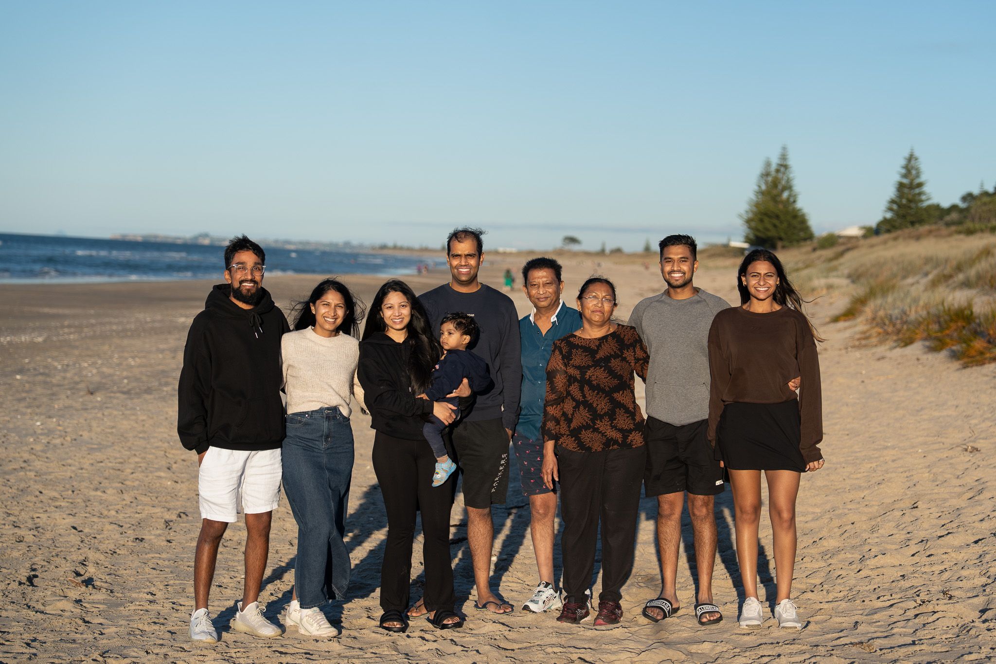 Extended family of nine standing together on a sandy beach, smiling towards the camera during a relaxed outdoor family portrait session in Auckland