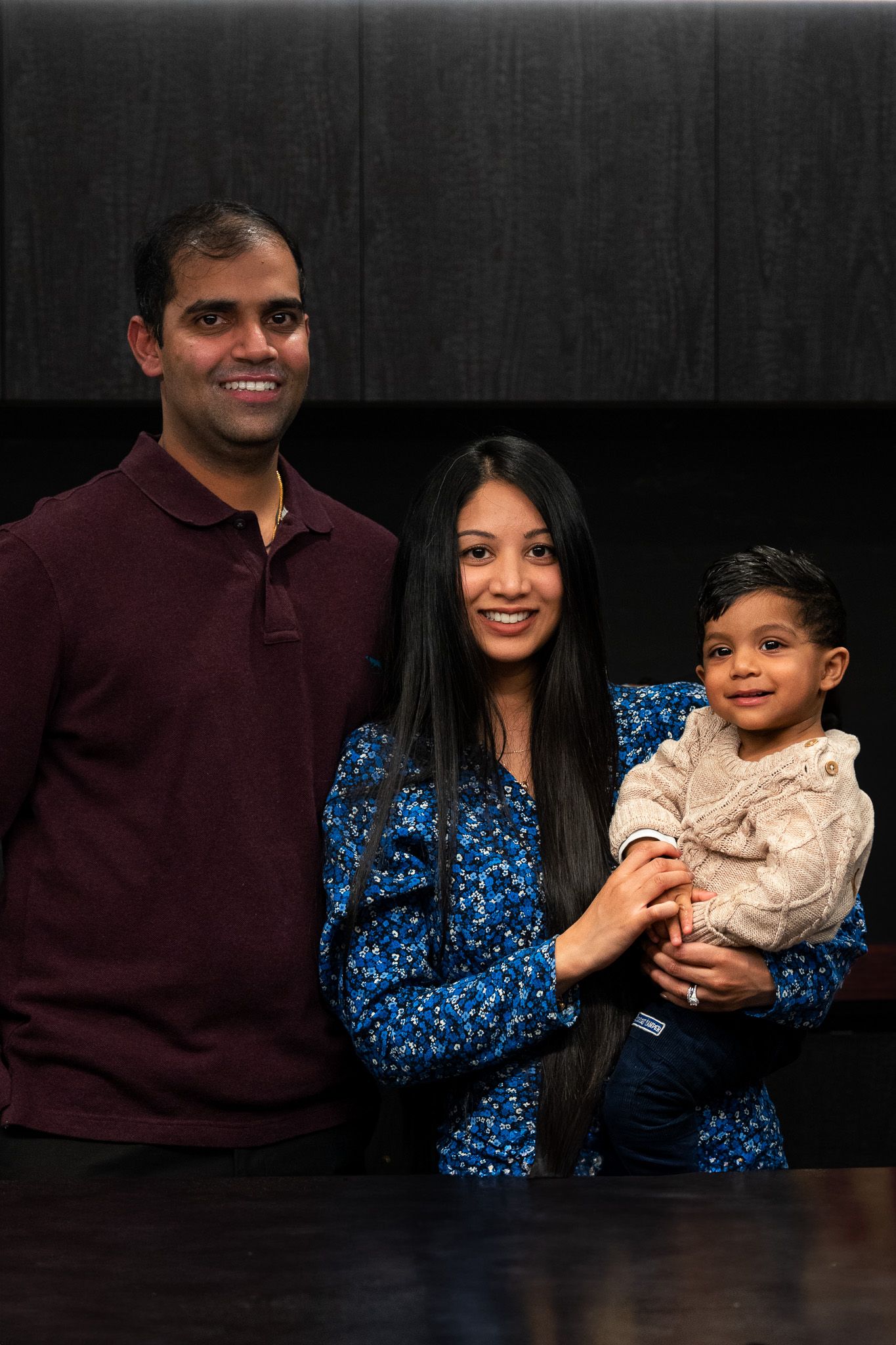 Young family of three posing indoors with parents smiling and holding their toddler during a warm and intimate family portrait session in Auckland