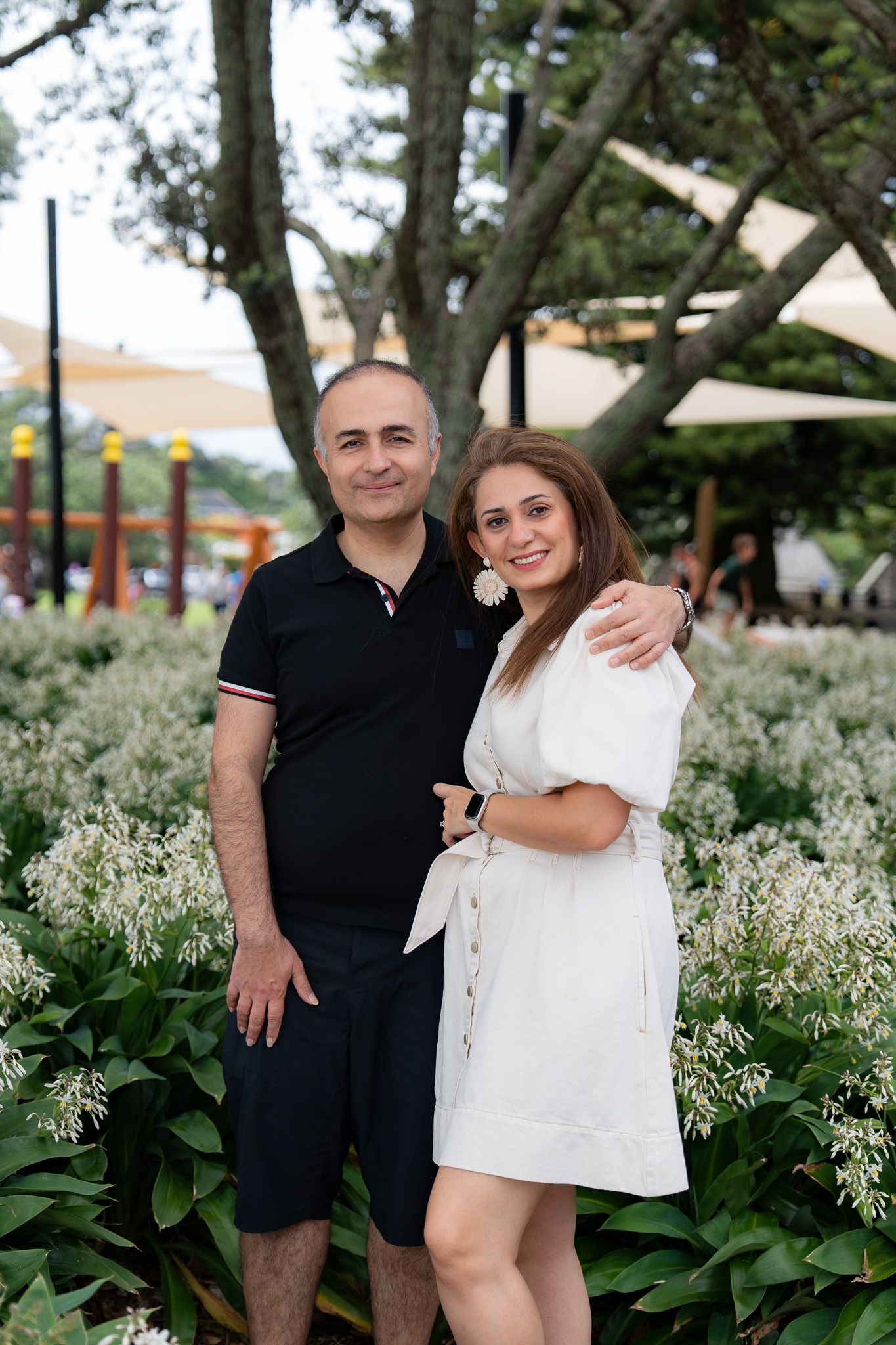 Couple smiling and posing together in a garden near Mission Bay Beach in Auckland during a family photography session surrounded by white flowering plants