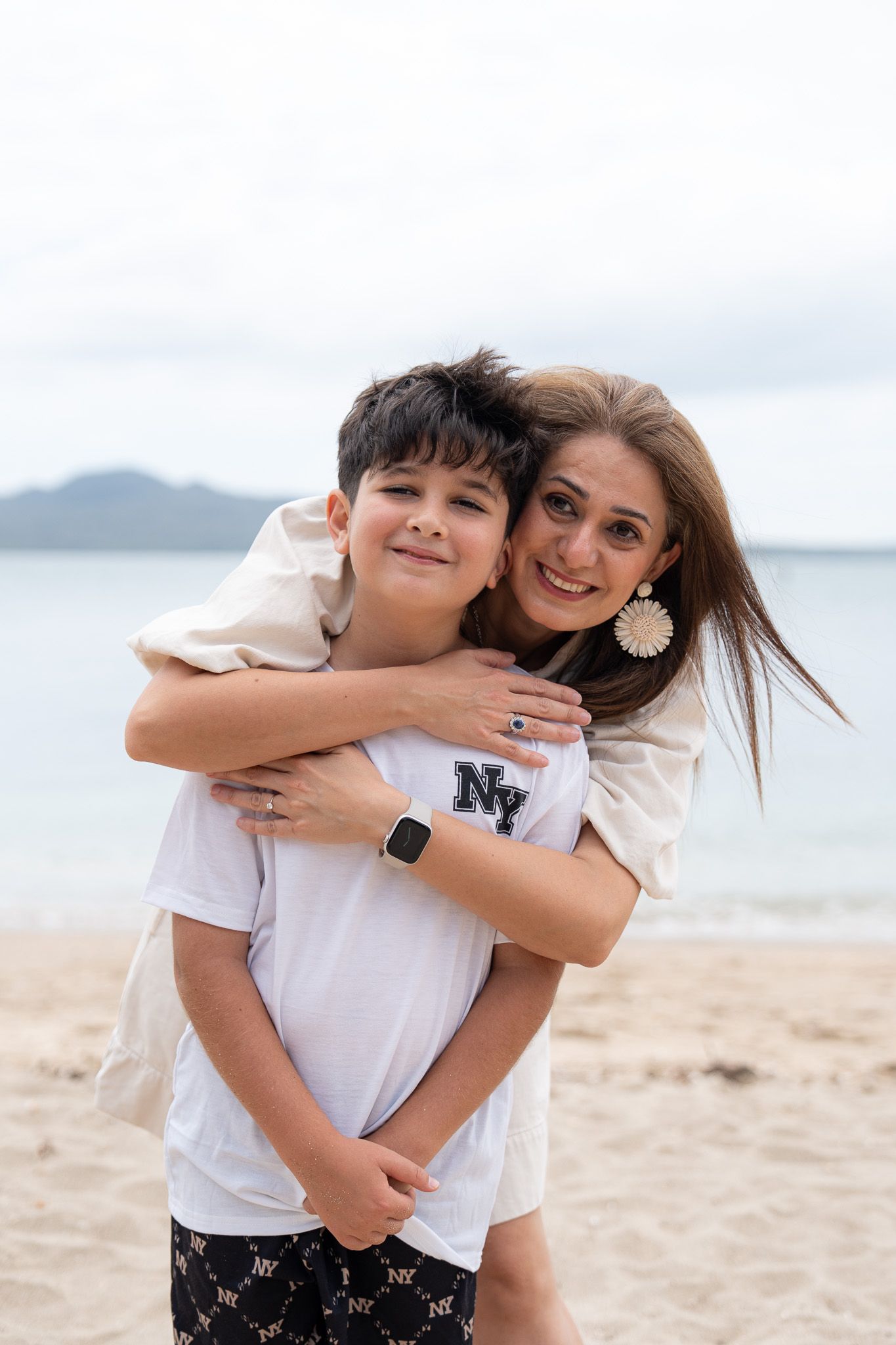 Mother hugging her son from behind during a warm family photography moment at Mission Bay Beach in Auckland with soft ocean and Rangitoto views behind them