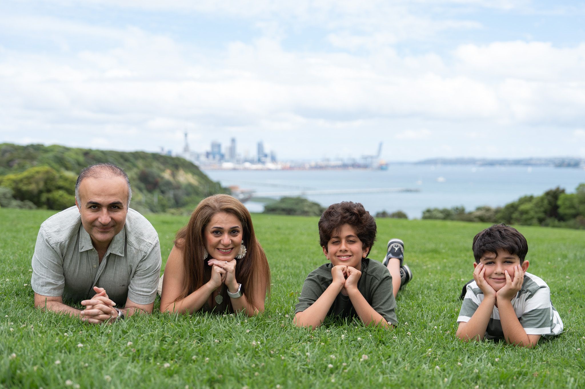 Family of four lying on the grass with Auckland city skyline and harbour behind them during a family photography session at the Michael Joseph Savage Memorial in Mission Bay