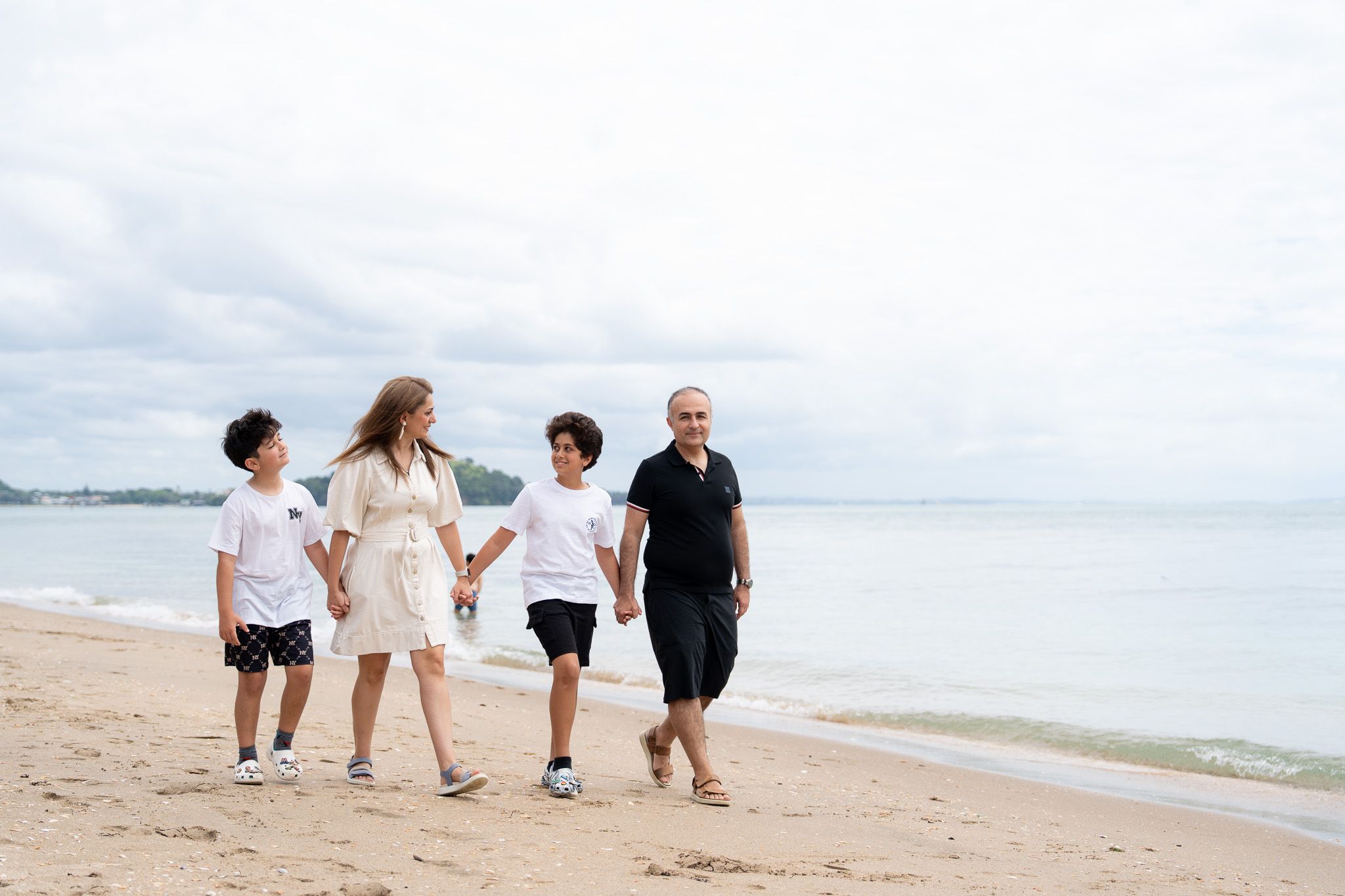 Family of four walking hand in hand along the shoreline during a relaxed family photography session at Mission Bay Beach in Auckland with calm water behind them