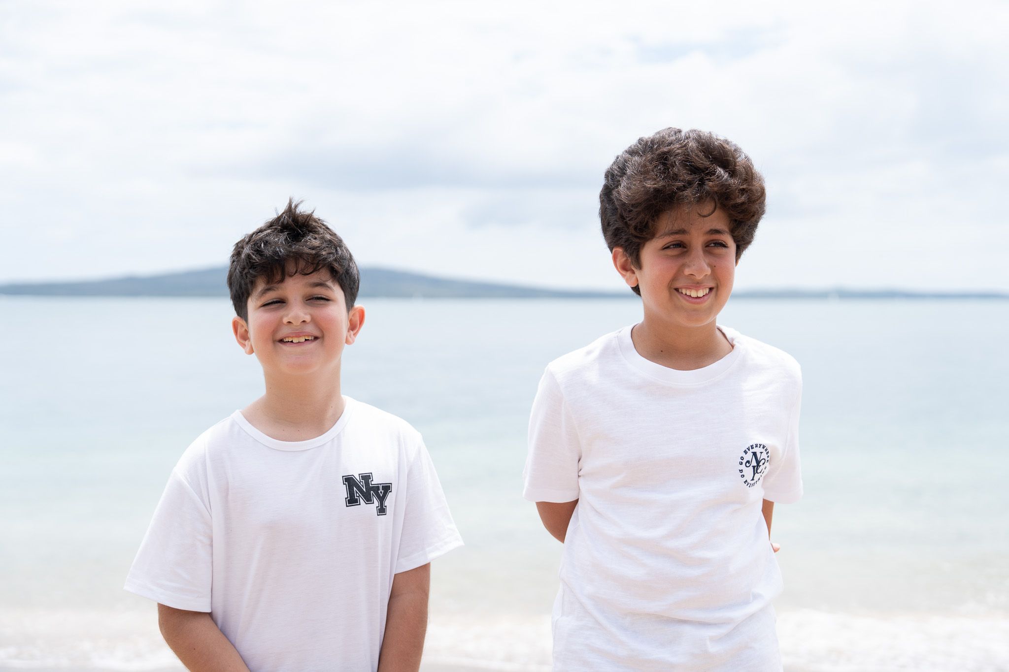 Two brothers smiling at the camera during a family photography session on Mission Bay Beach in Auckland with Rangitoto Island visible in the background