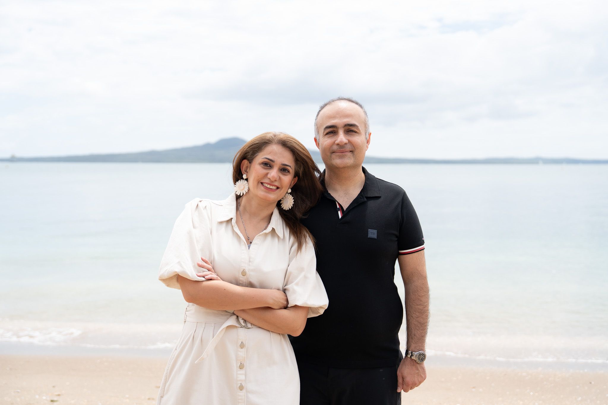 Couple smiling together during a beachside family photography session at Mission Bay in Auckland with calm ocean water and Rangitoto Island behind them