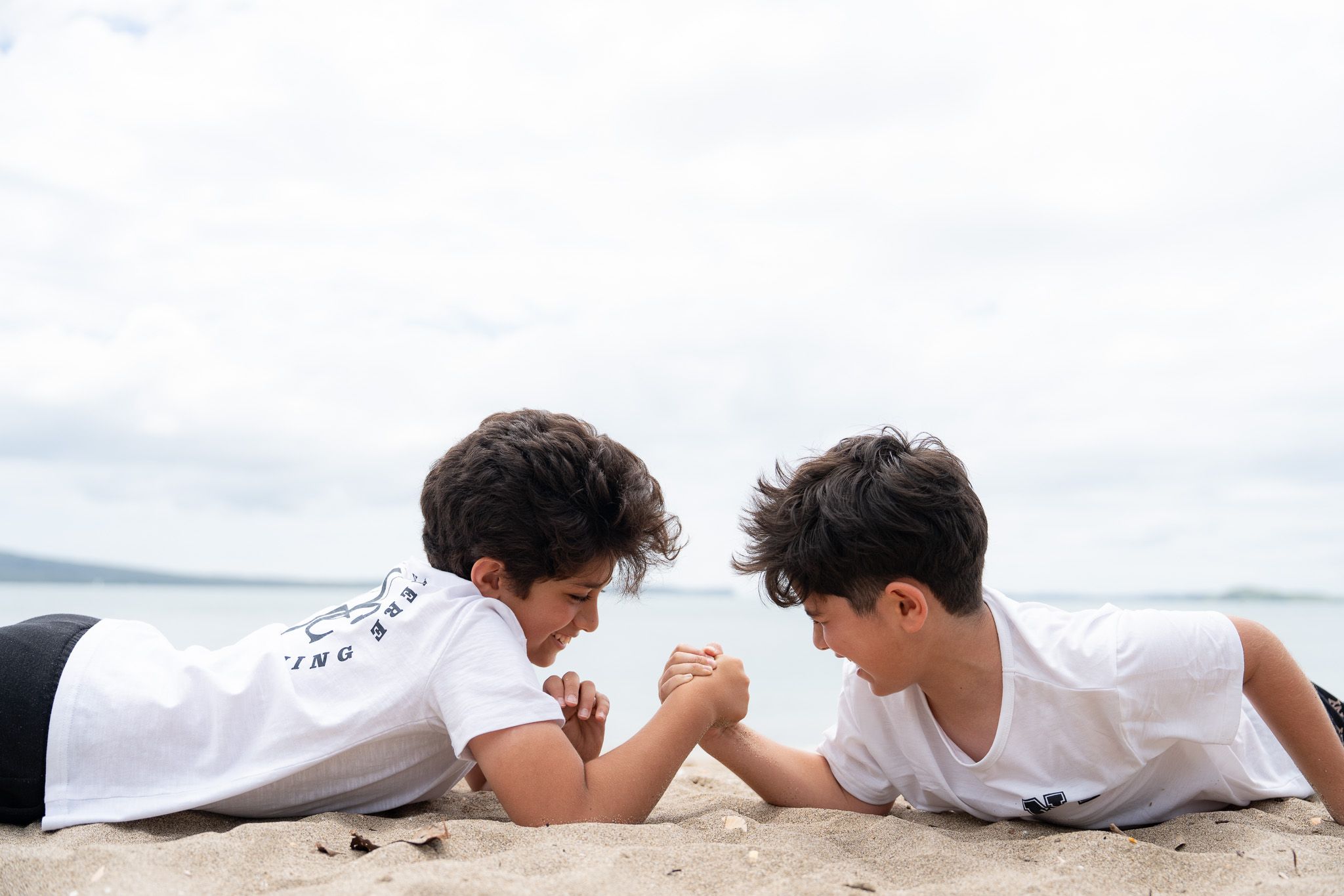 Two brothers laughing and arm-wrestling on the sand during a fun family photography session at Mission Bay Beach in Auckland with the ocean behind them