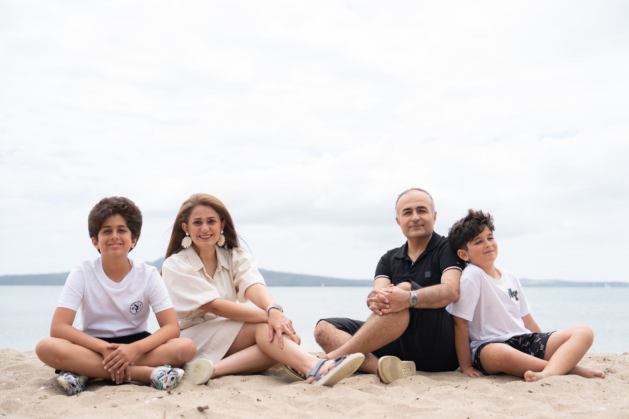Family of four sitting on the sand during a relaxed family photography session at Mission Bay Beach in Auckland with calm water and Rangitoto Island in the distance