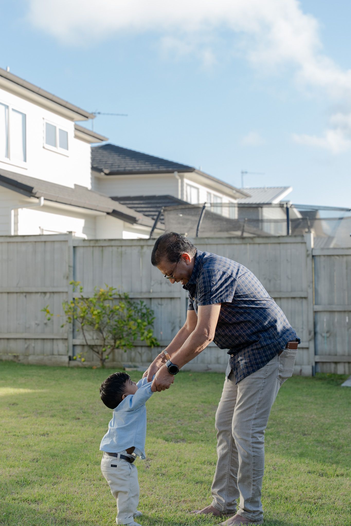 Grandfather playing with his young grandson in the backyard, holding his hands and spinning him around during a candid family session in Auckland