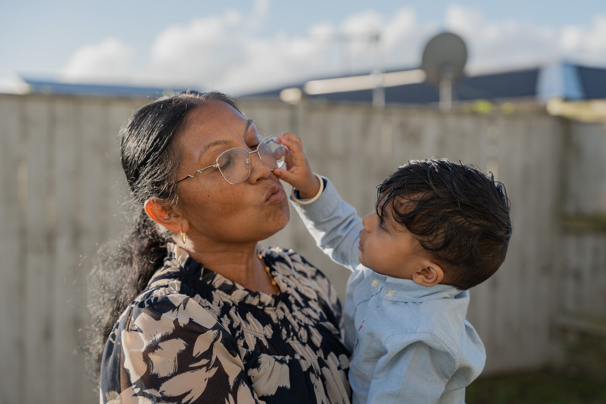 Grandmother smiling as her young grandson reaches up to touch her glasses during a playful candid family moment in Auckland