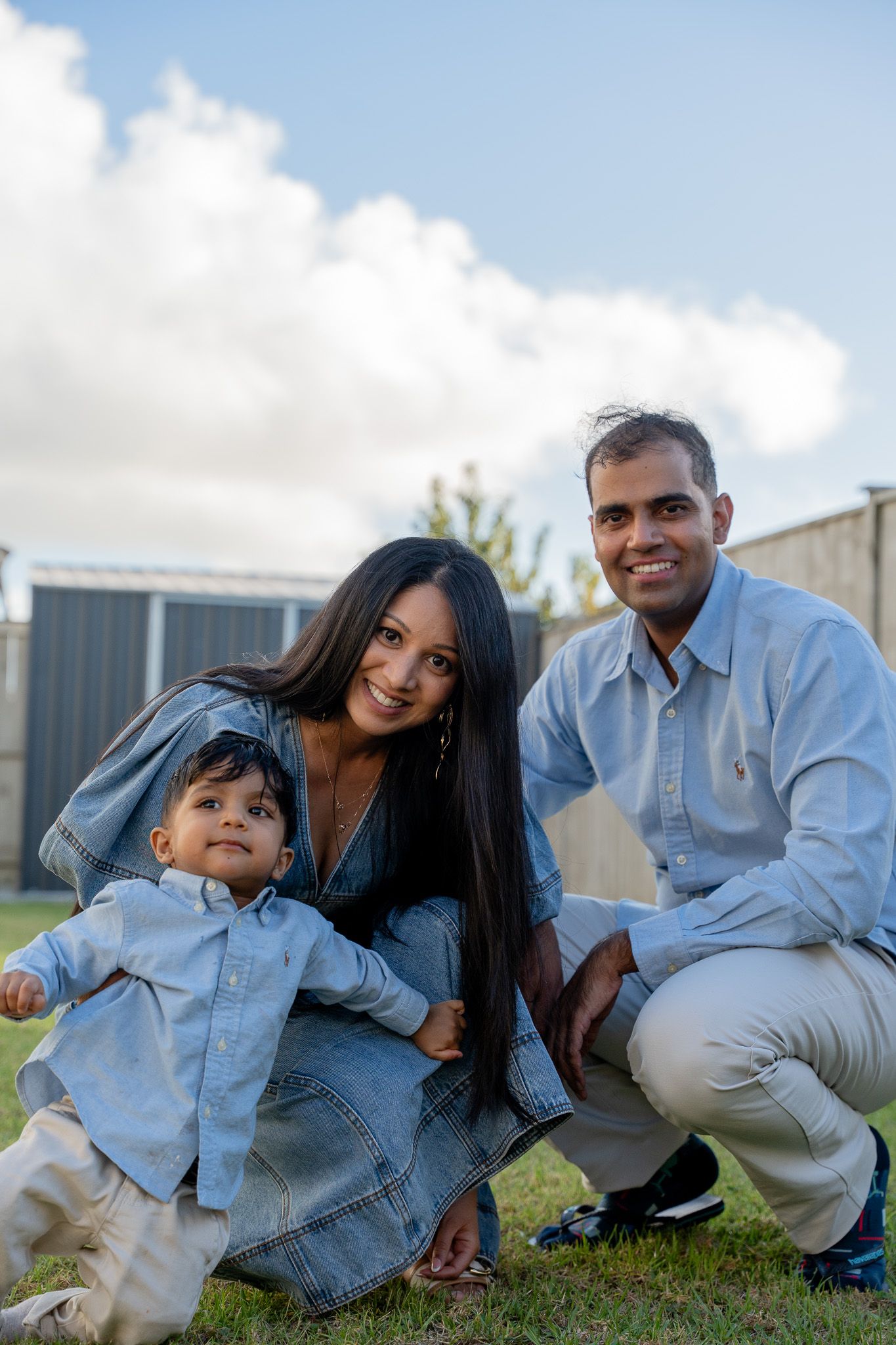 Family of three crouching together in the backyard, smiling at the camera during a relaxed outdoor family photoshoot in Auckland