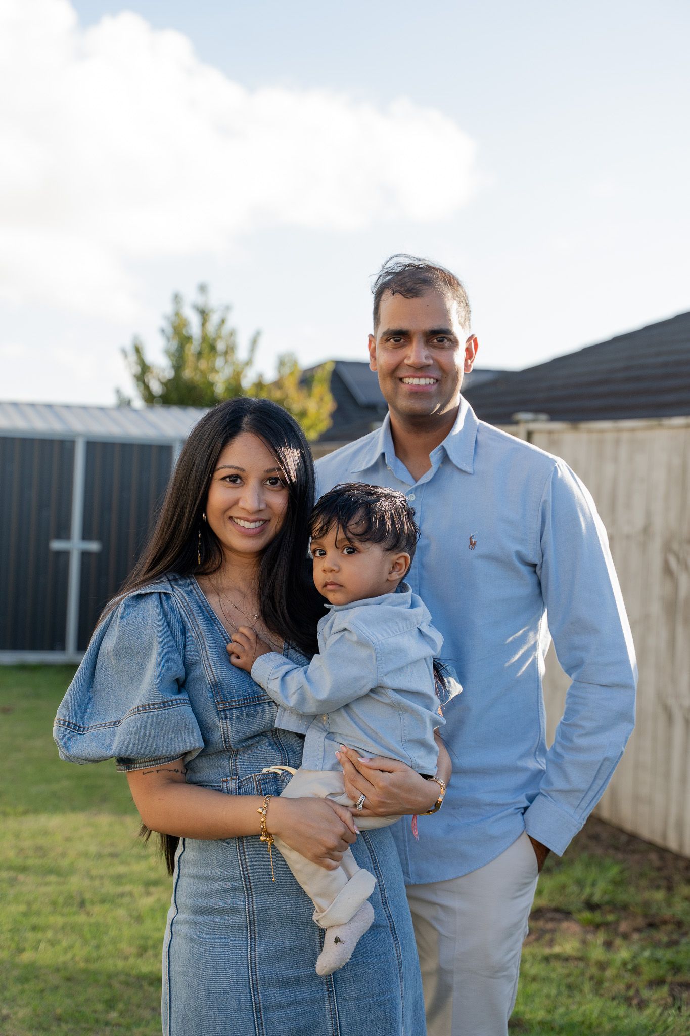Young family of three posing in the backyard with their toddler in the mother’s arms during a golden hour outdoor family session in Auckland