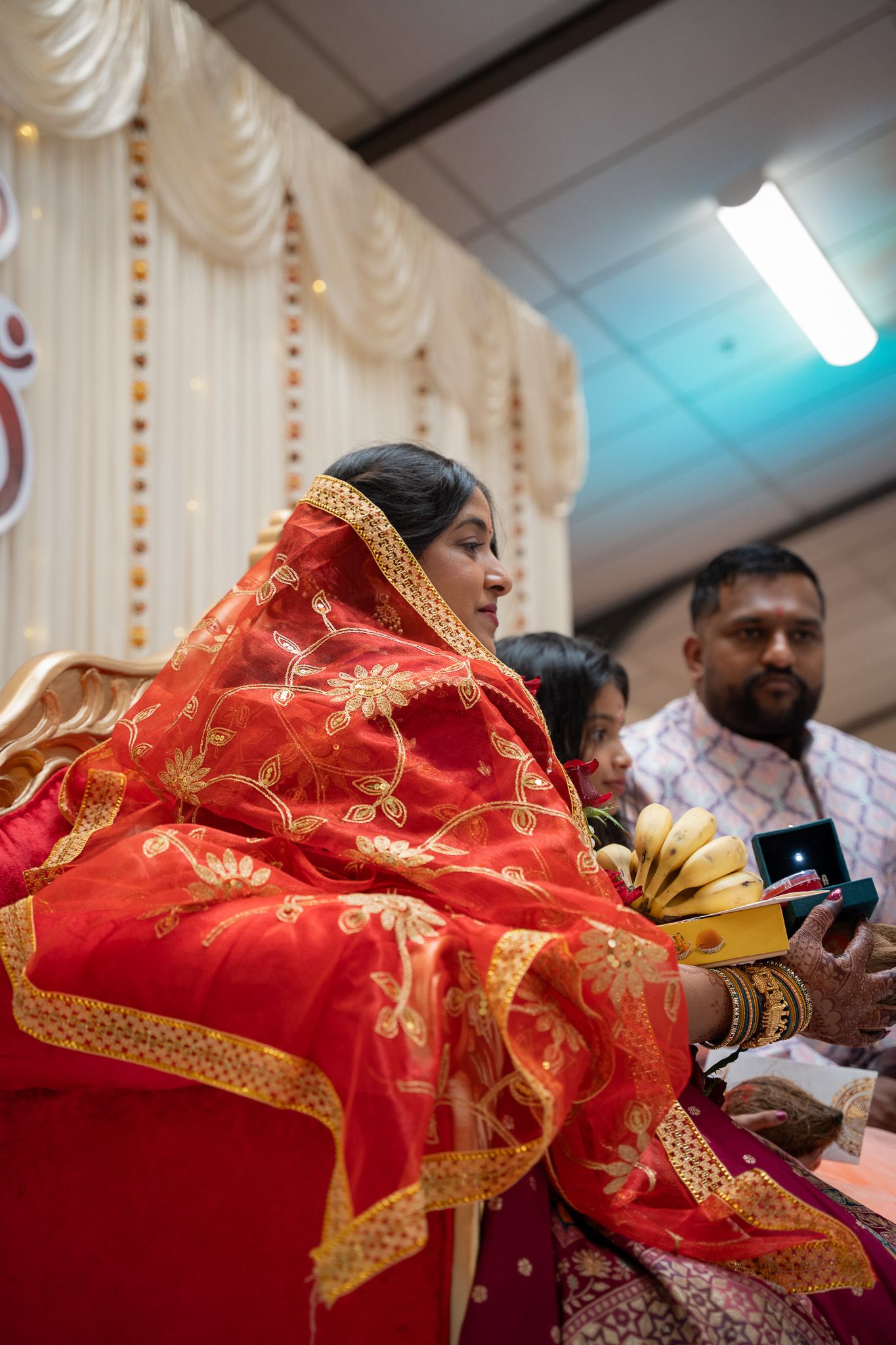 Bride draped in red embroidered dupatta holding ceremonial offerings with groom seated beside her during engagement in Auckland