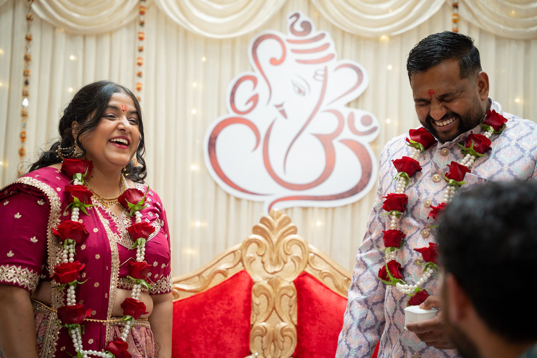 Couple laughing together on decorated stage wearing rose garlands during joyful Indian engagement celebration in Auckland