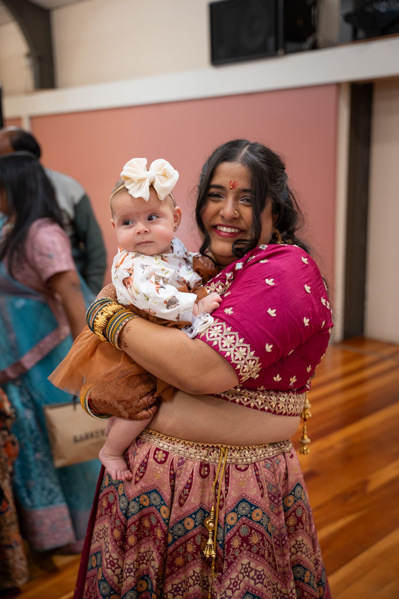 Bride holding smiling baby dressed in white and brown outfit during Indian engagement celebration in Auckland