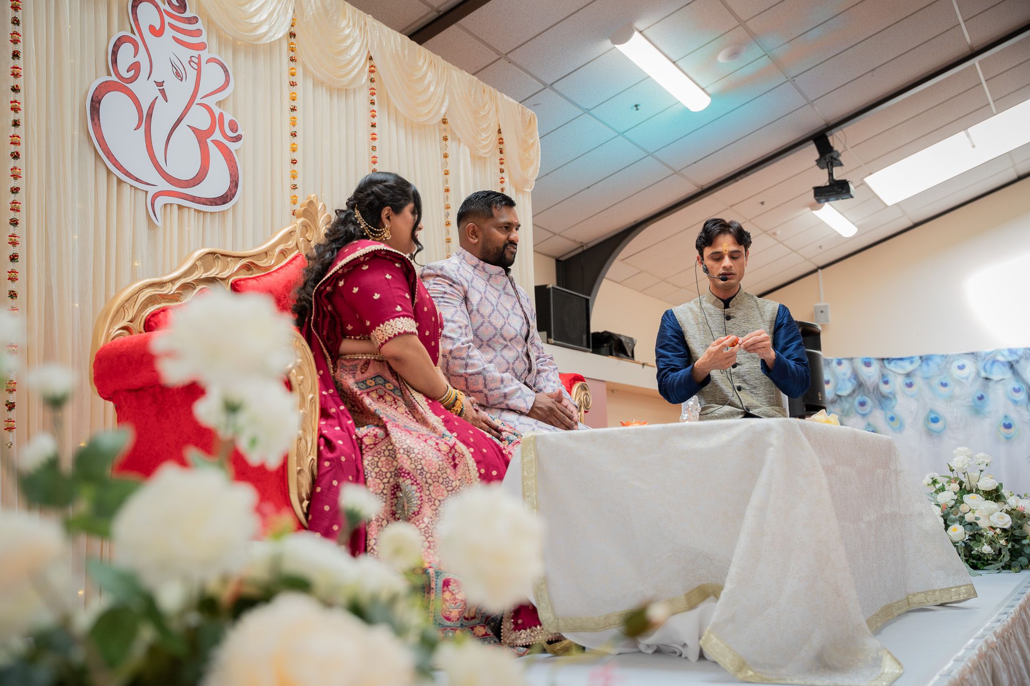 Bride and groom seated on stage listening to officiant during Indian engagement ceremony with floral decor in Auckland