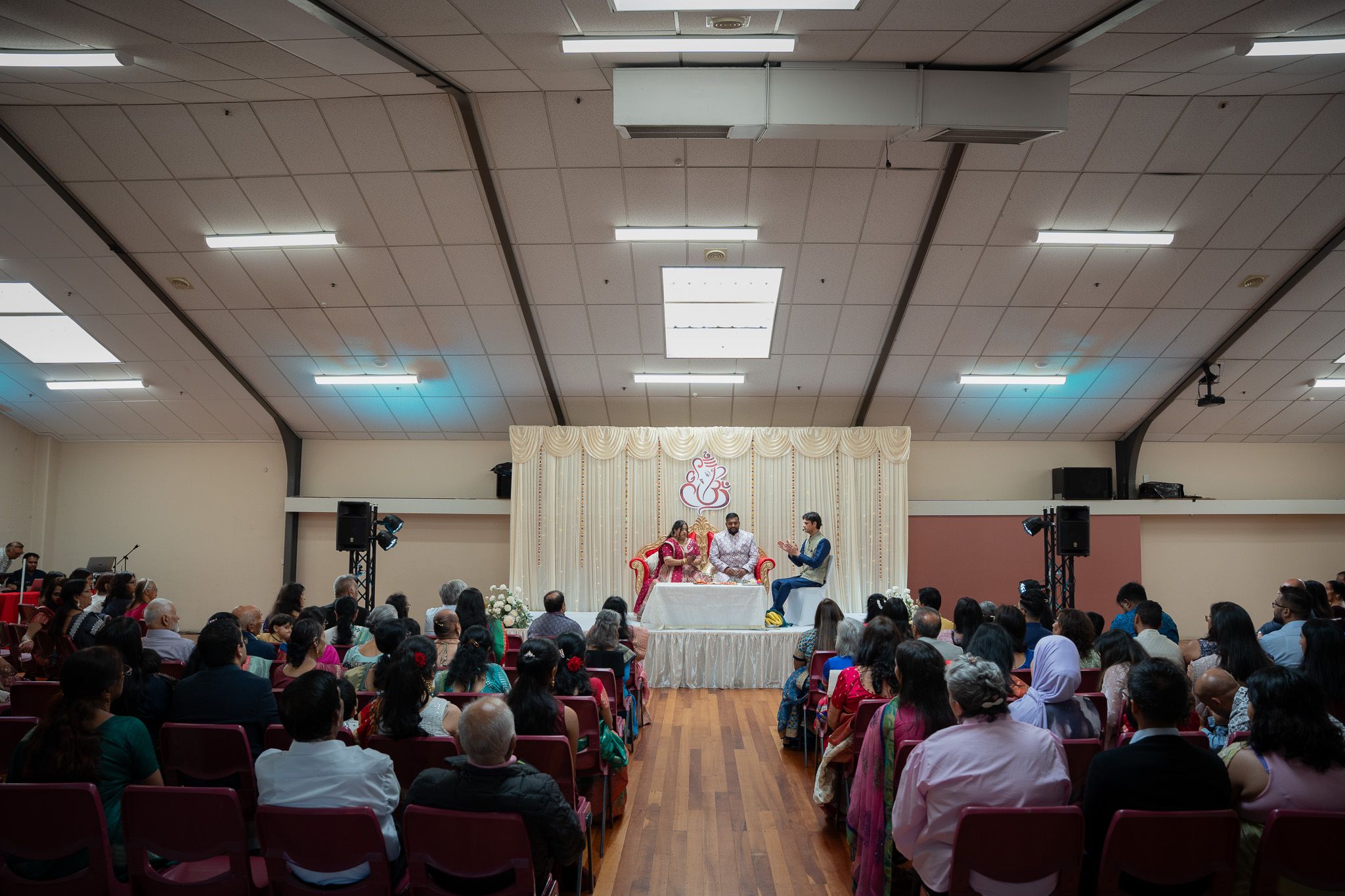 Wide view of decorated stage with bride, groom and officiant during Indian engagement ceremony in Auckland surrounded by guests
