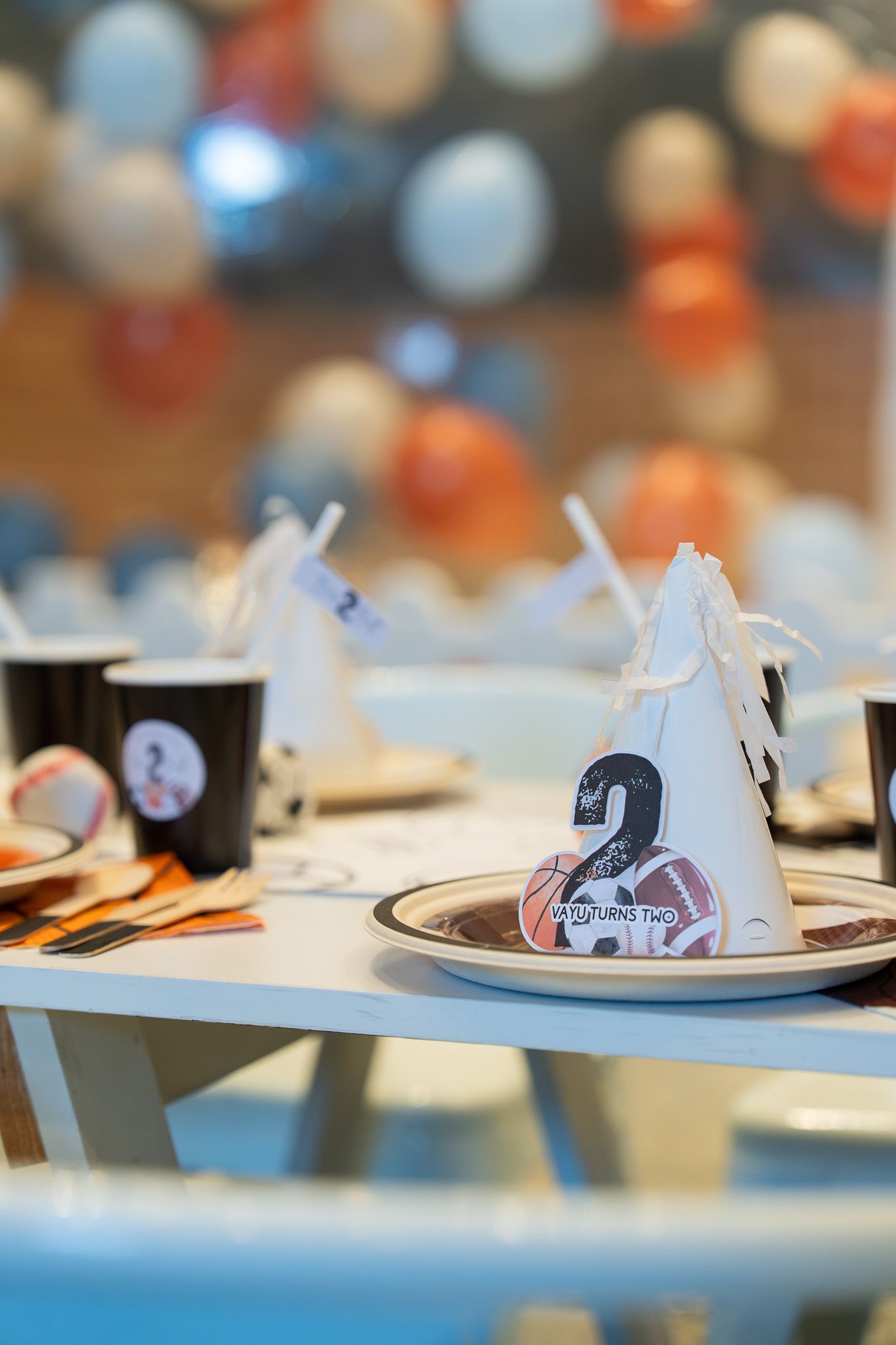 Close up of Vayu turns two party hat and sports themed table decor with balloons softly blurred in the background