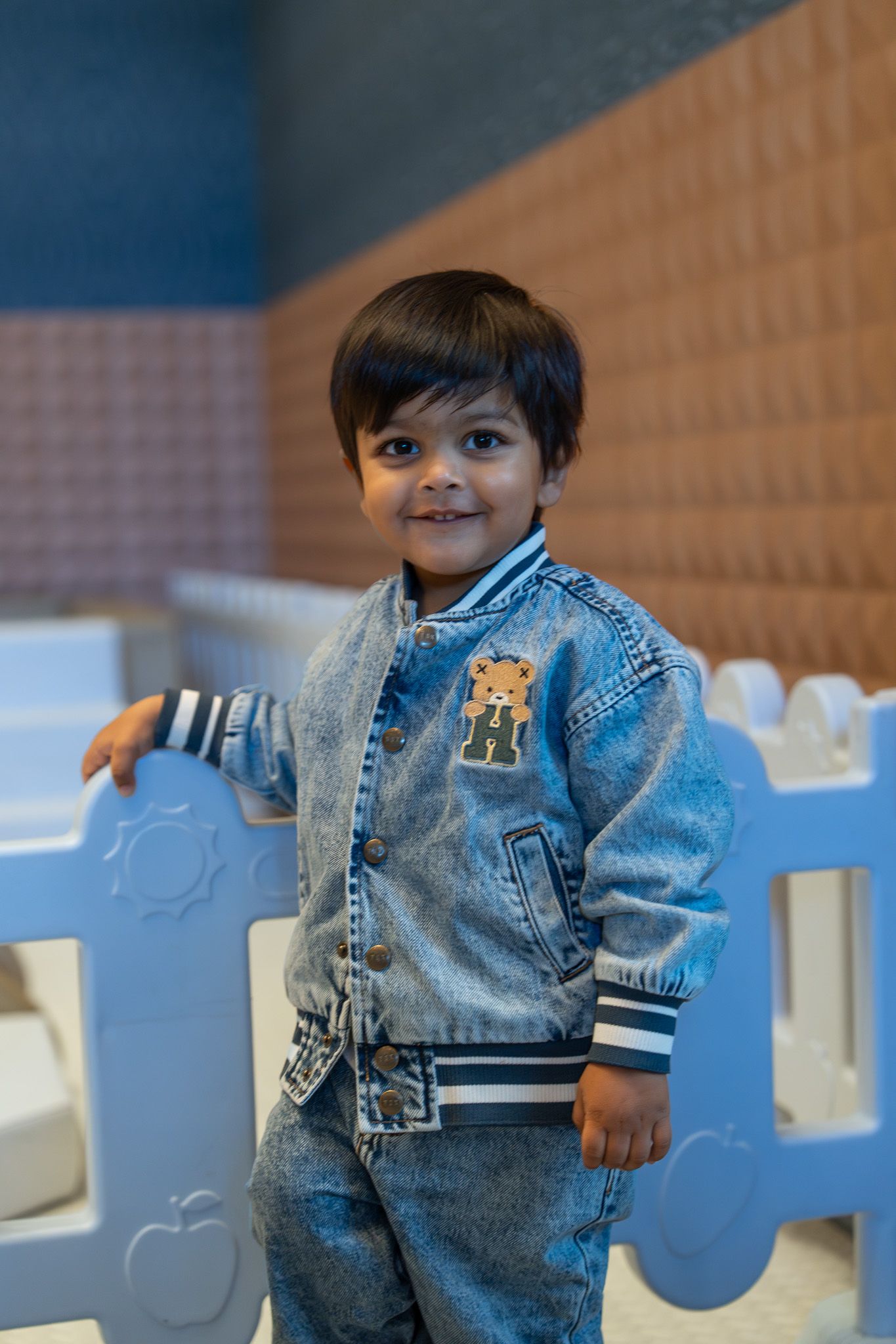 Smiling toddler in denim outfit standing by a white play fence during his 2nd birthday party at Naumi Hotel in Auckland
